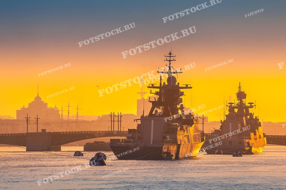 На этой фотографии изображен красочный рассвет или закат в Санкт-Петербурге, где небо и река Нева окрашены в яркие оранжевые оттенки. Военные корабли и силуэты зданий создают драматичную и величественную картину. Такой снимок может быть использован в качестве иллюстрации к статьям о Военно-морском флоте, в фотографических коллекциях с видами городов, или для дизайна интерьеров, где хочется добавить элементы морской эстетики. Также он прекрасно подойдёт для публикаций о путешествиях и культурных мероприятиях, проходящих в городе.